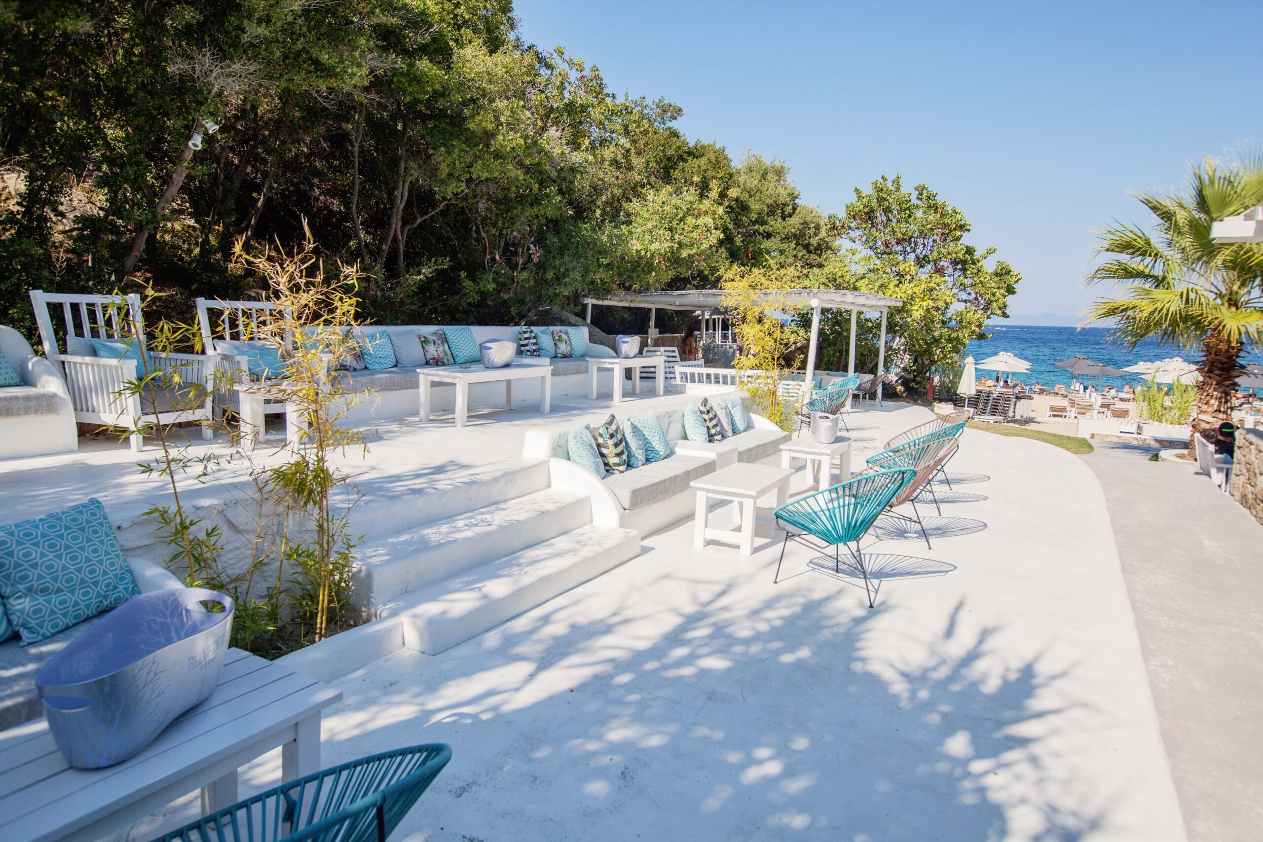 Outdoor lounge area with white steps, blue chairs, cushioned benches, and potted plants overlooking a beach with sunbeds and umbrellas, surrounded by greenery and ocean views.