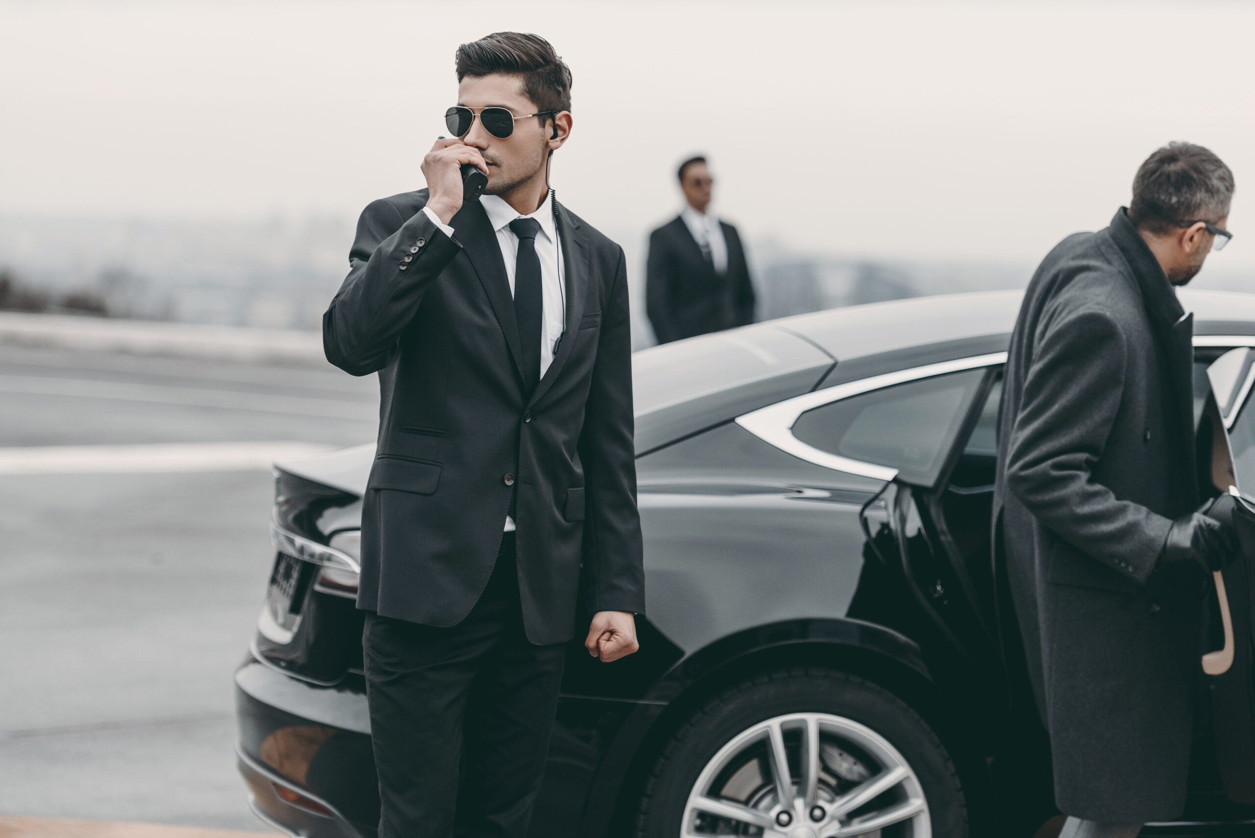 Three men in suits stand near a black car. One man in sunglasses speaks into a walkie-talkie, another stands in the background, and the third is opening the car door, suggesting a security or bodyguard scenario.
