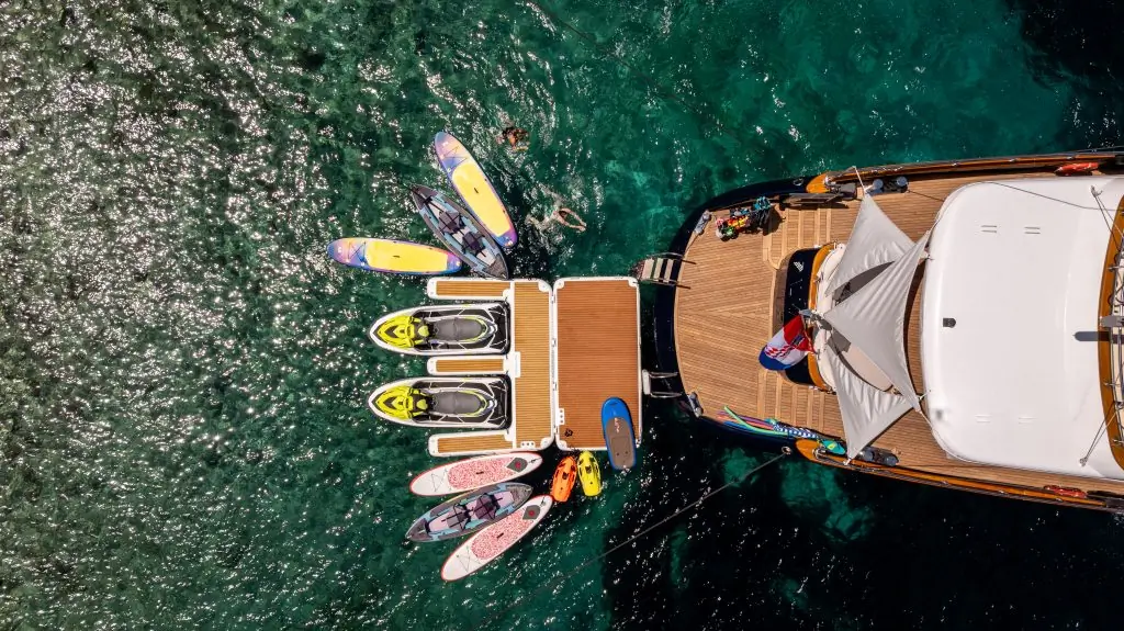 Aerial view of a yacht docked on clear blue water with paddleboards, jet skis, and kayaks lined up at the back. Several people are swimming and enjoying water activities around the boat.