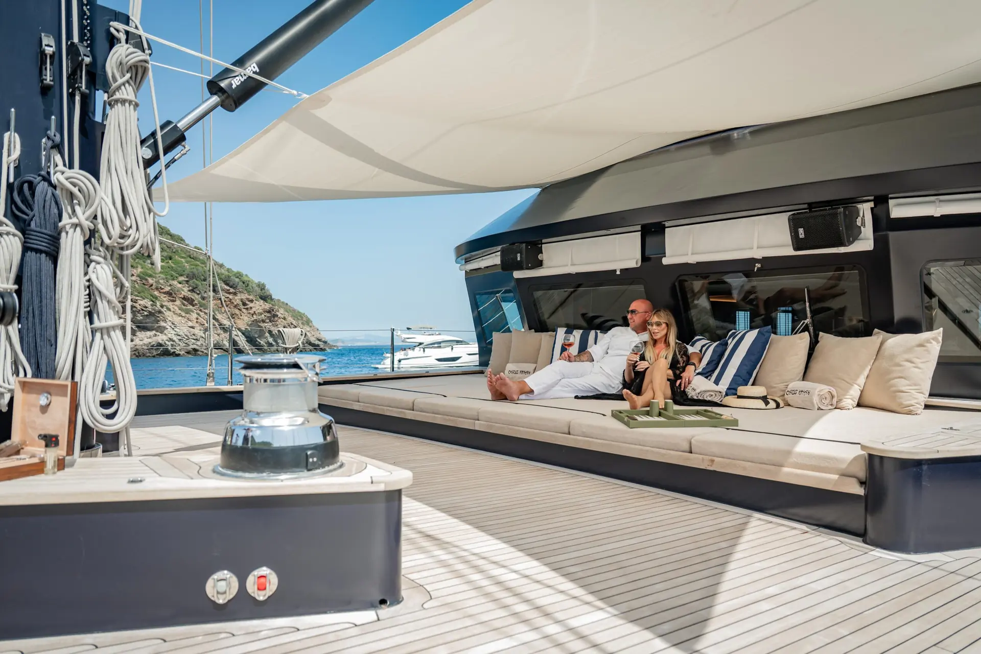 A man and woman relax on a large yacht deck under a canopy, with blue water and another boat visible in the background on a sunny day.
