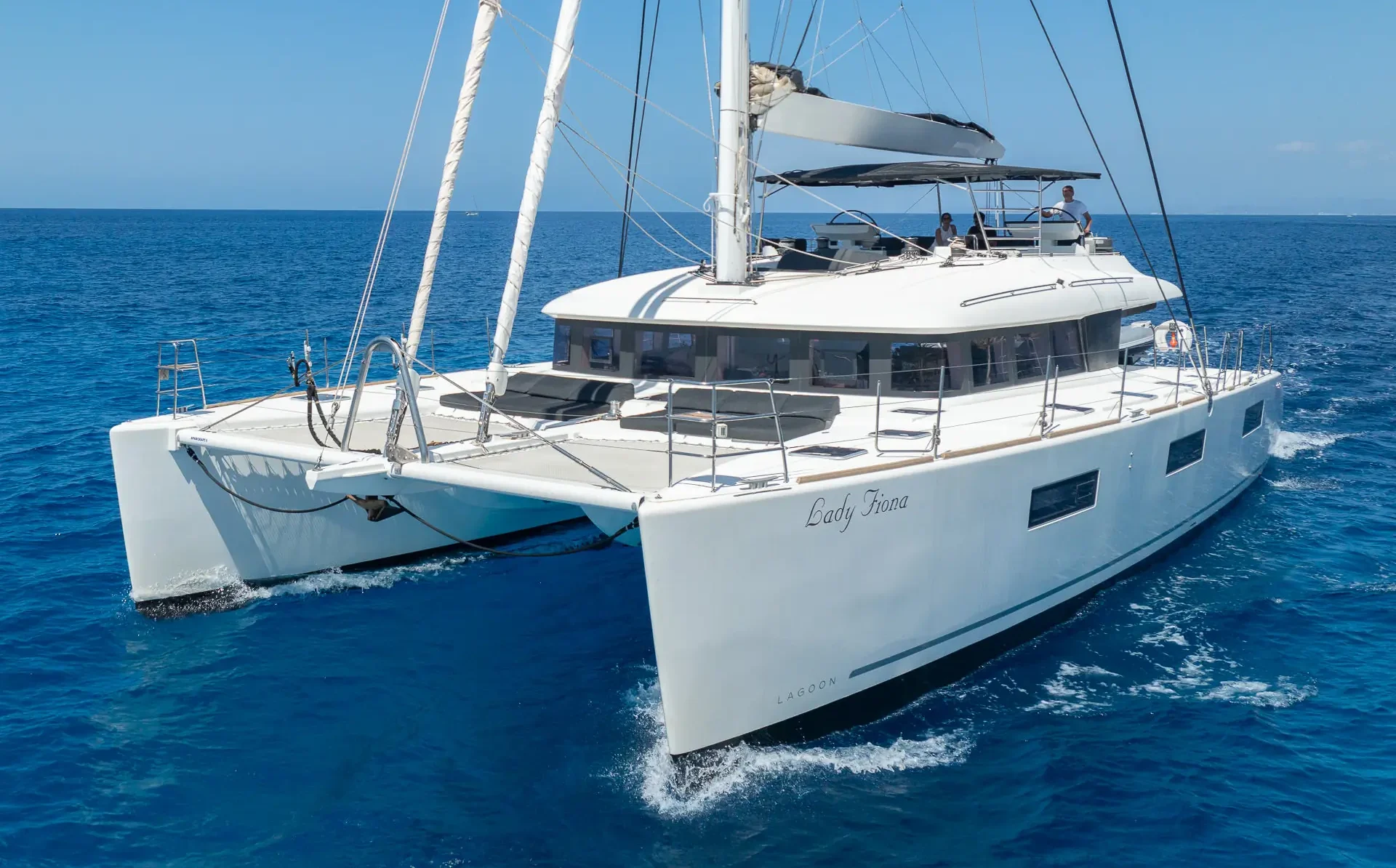 A large white catamaran yacht sails on clear blue ocean water under a sunny sky. The yacht has two hulls, multiple decks, and the name “Lady Fiona” written on the side.