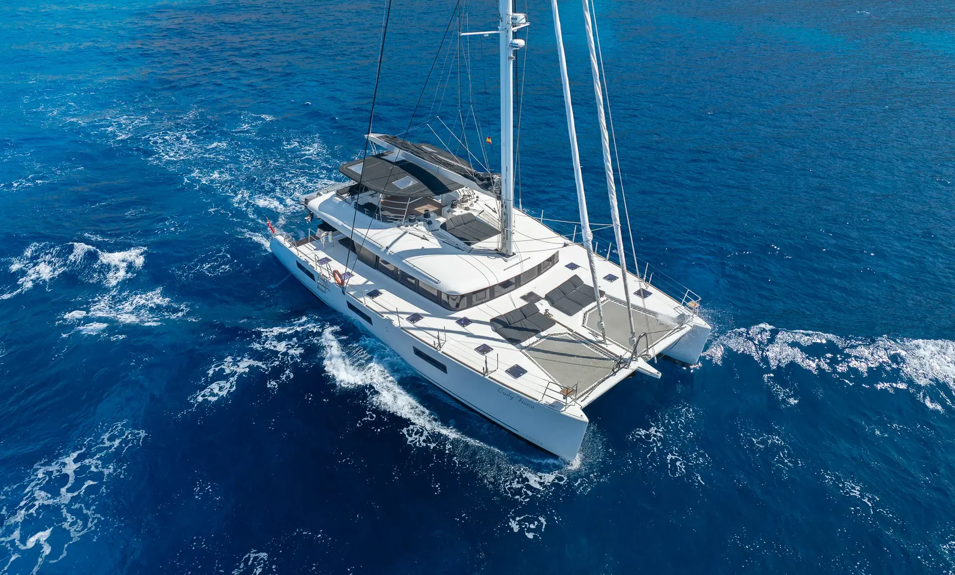 A white catamaran sails on clear blue water near a rocky coastline with green vegetation under a sunny sky.