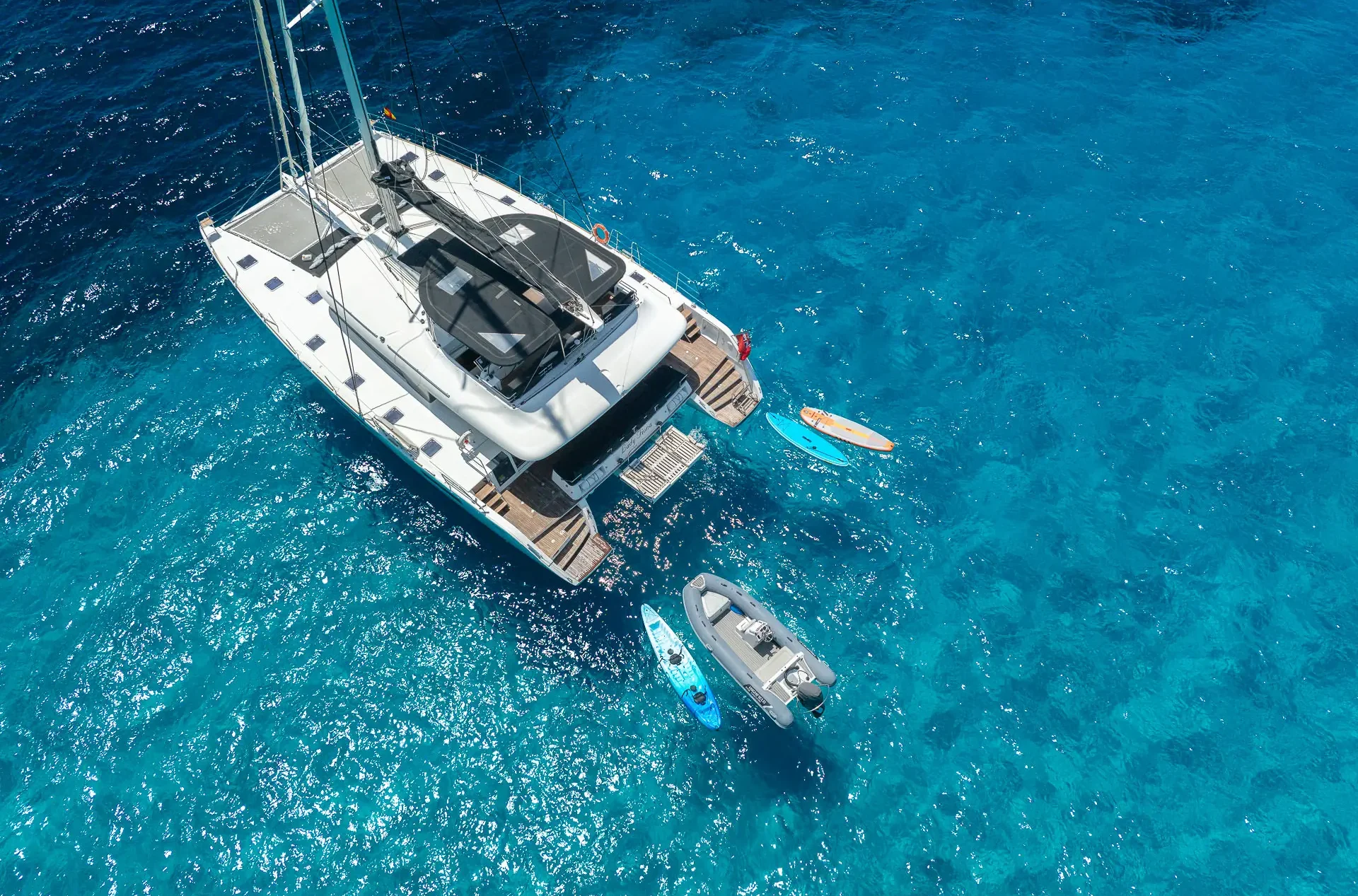 Aerial view of a white catamaran yacht anchored in clear blue water, with a small dinghy and two paddleboards floating nearby.