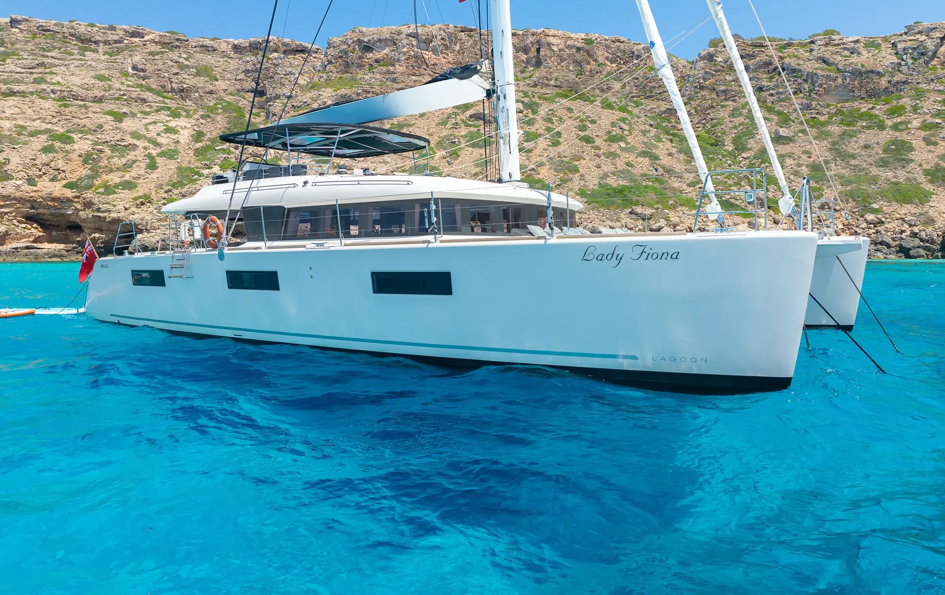 A white catamaran named Lady Fiona floats on clear blue water near rocky, shrub-covered cliffs under a bright blue sky.