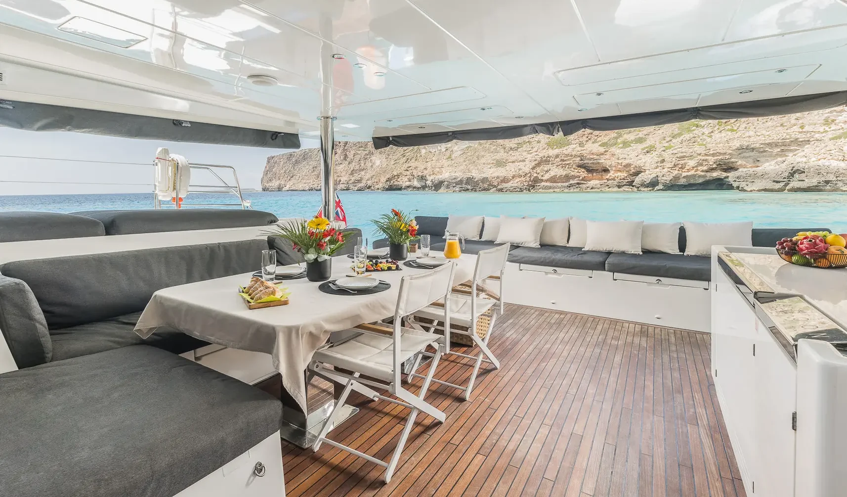 A luxurious yacht deck with a dining table set for a meal, cushioned seating, and a view of clear turquoise water and rocky coastline in the background.