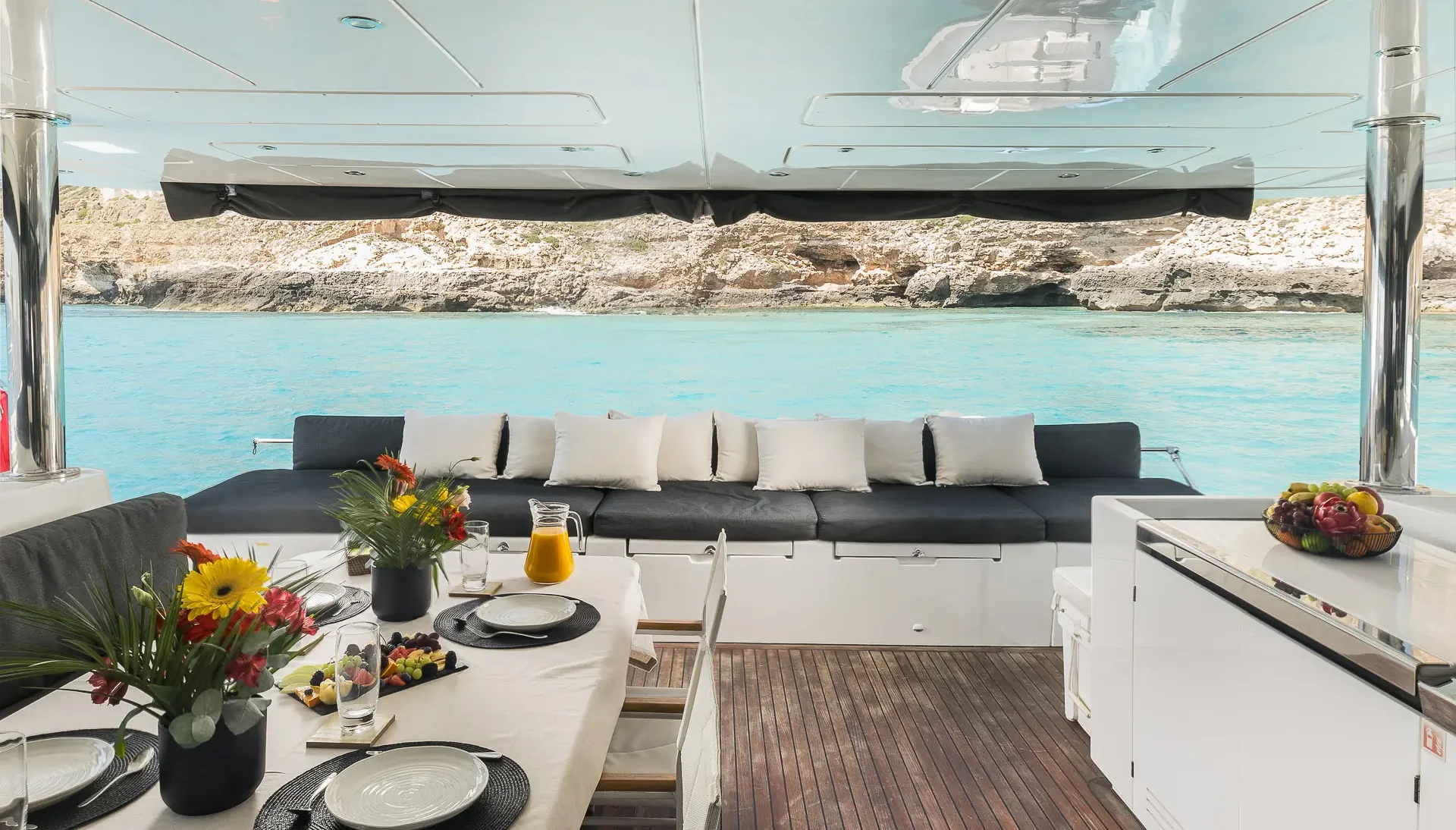 A yacht’s dining area with a table set for four, floral arrangements, fruit, and juice. Behind is a cushioned bench with pillows and a view of turquoise water and rocky shoreline through open windows.