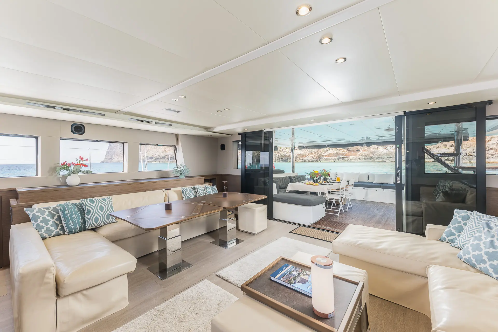 Luxurious yacht interior with beige leather sofas, wooden tables, large windows, and ocean views. Glass doors open to an outdoor seating area with a dining table, decorated with flowers and surrounded by natural light.