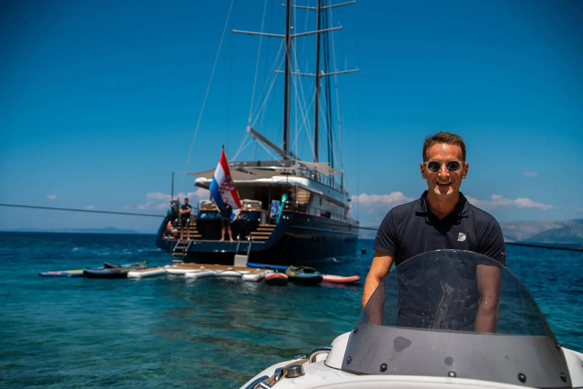 A man in sunglasses smiles on a small boat in clear blue water, with a large yacht and people in the background. The sky is clear and sunny.