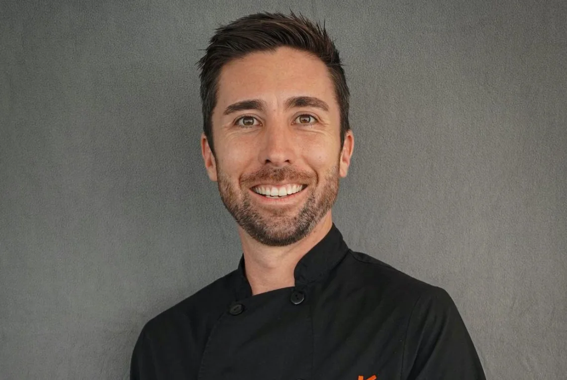 A smiling man with short brown hair and a trimmed beard wears a black chefs jacket and stands against a plain gray background.