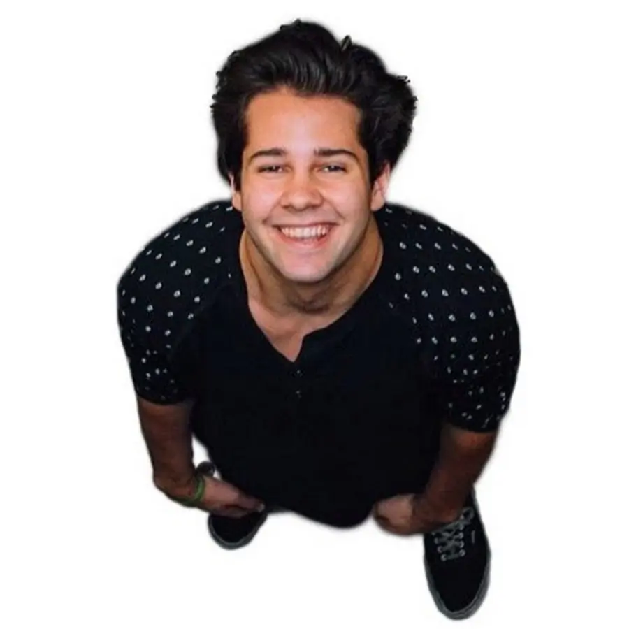 A young man with dark hair smiles, looking up at the camera. He wears a black shirt with patterned sleeves and black shoes, standing against a white background.