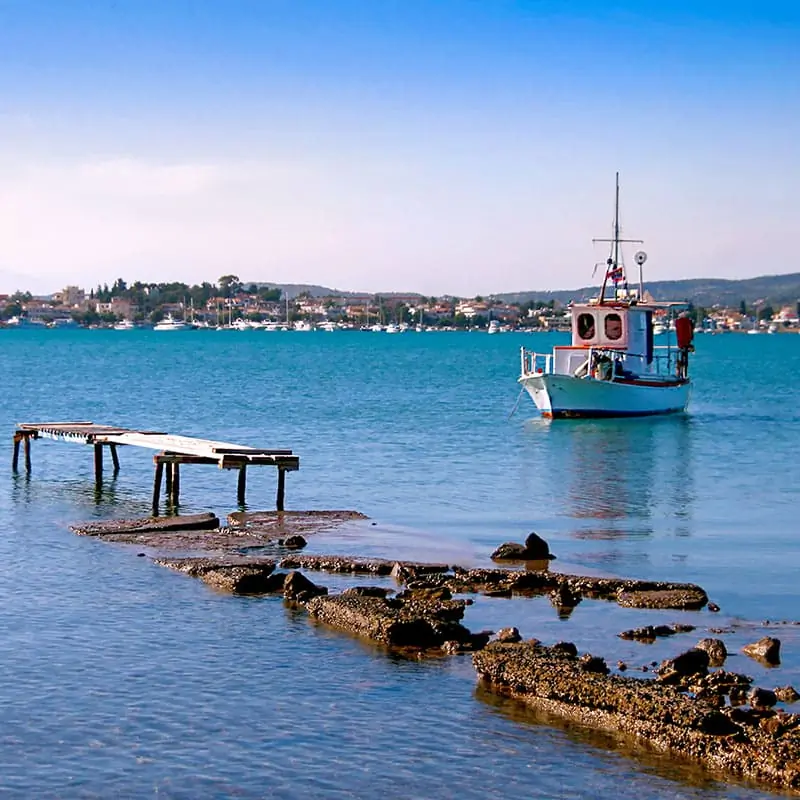 Destroyed pier and white small wooden fishing boat. View of Porto Heli town in the background. Greece