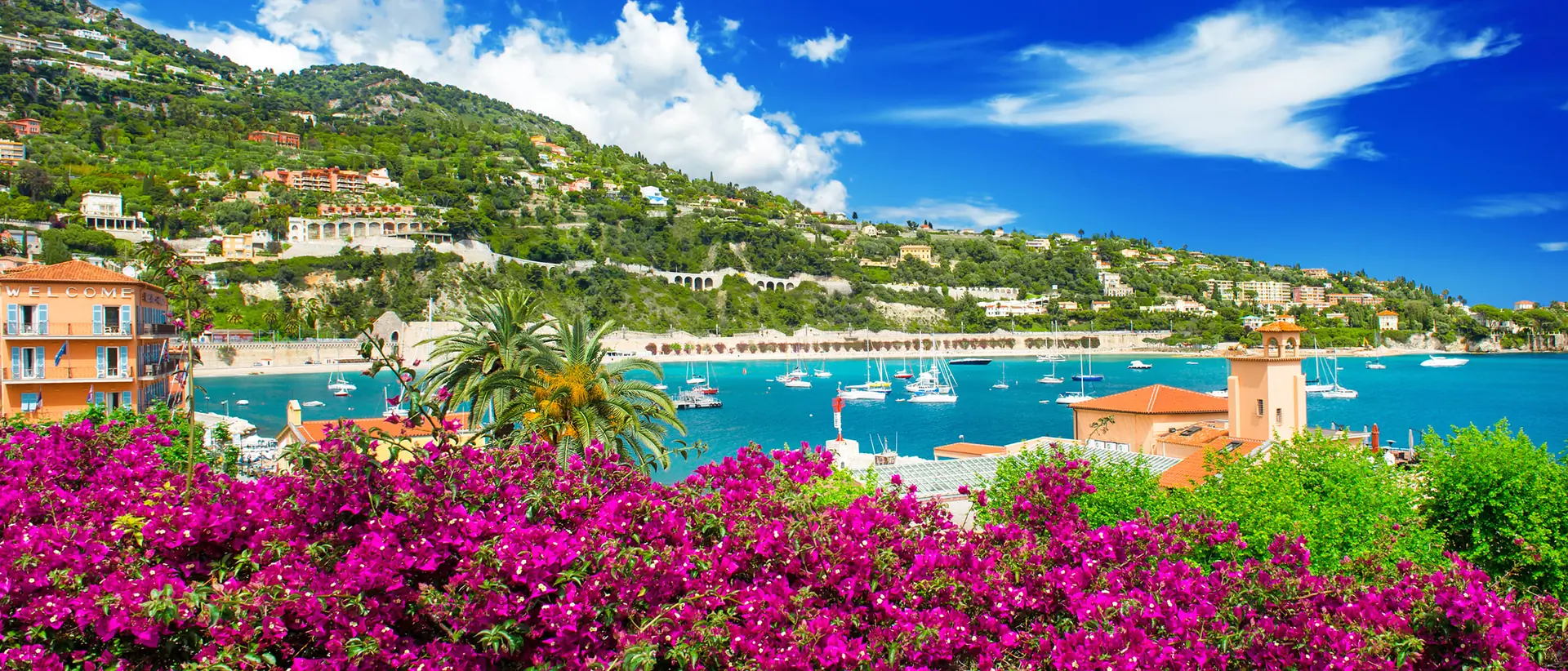 Colorful coastal town with bright pink bougainvillea, turquoise water dotted with boats, and green hills with houses under a blue sky with clouds.