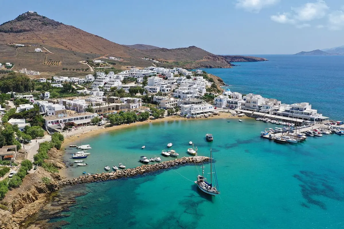Aerial view of a coastal village with white buildings, clear turquoise water, boats in a small harbor, and hills in the background under a blue sky.