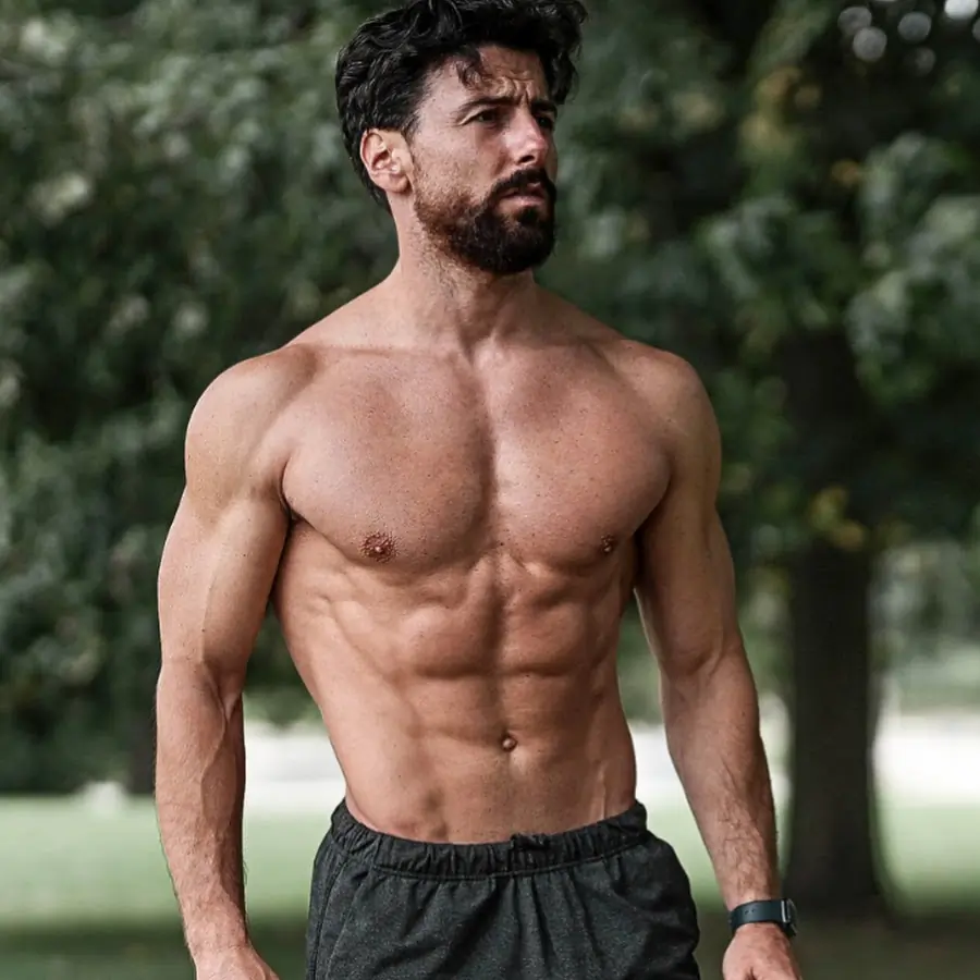 A muscular, shirtless man with dark hair and a beard stands outdoors in front of trees, wearing dark athletic shorts and a fitness tracker on his wrist.