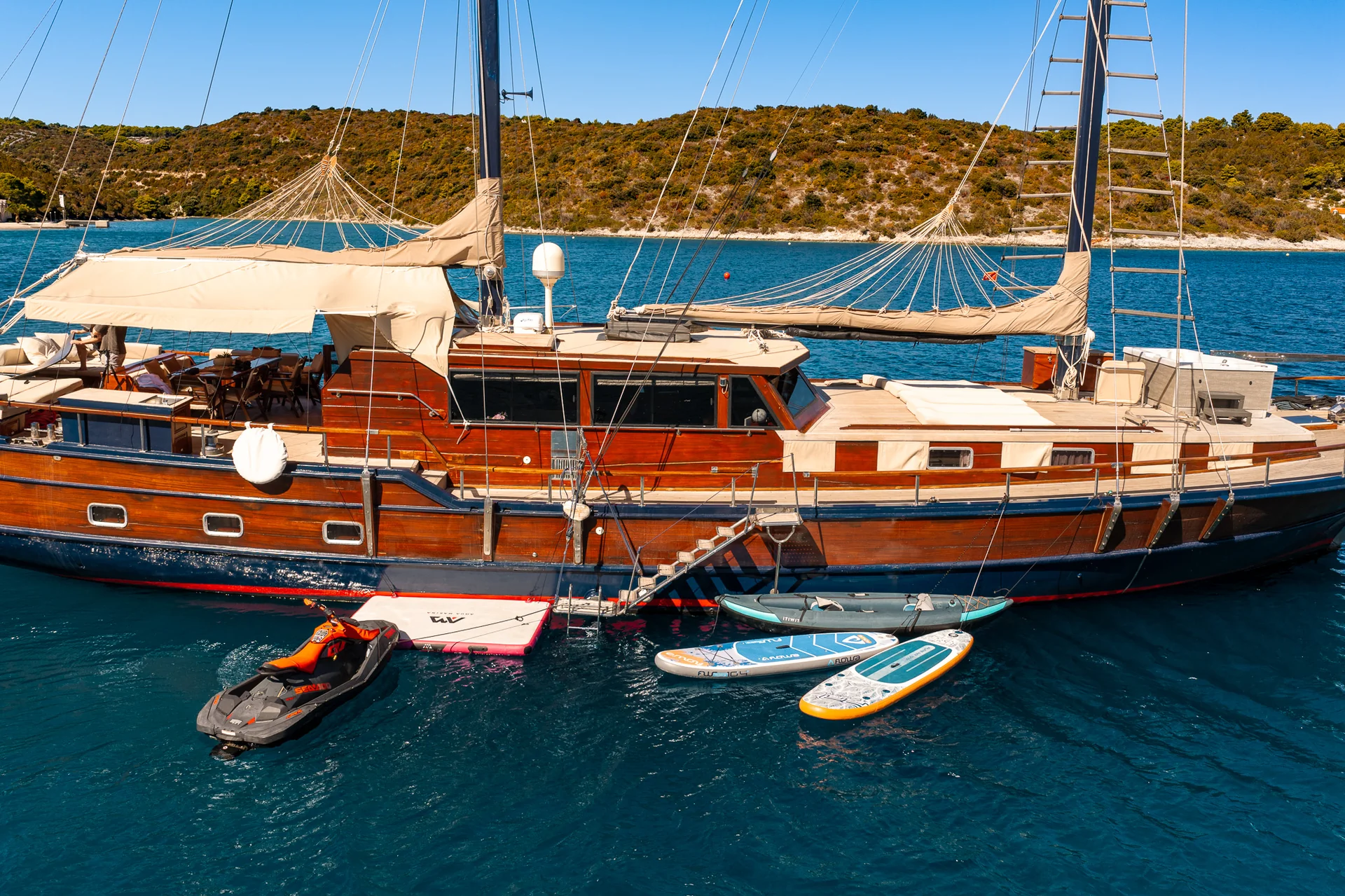 PACHA A wooden motor sailer anchored in blue water with a jet ski, an inflatable platform, and three paddleboards moored alongside.