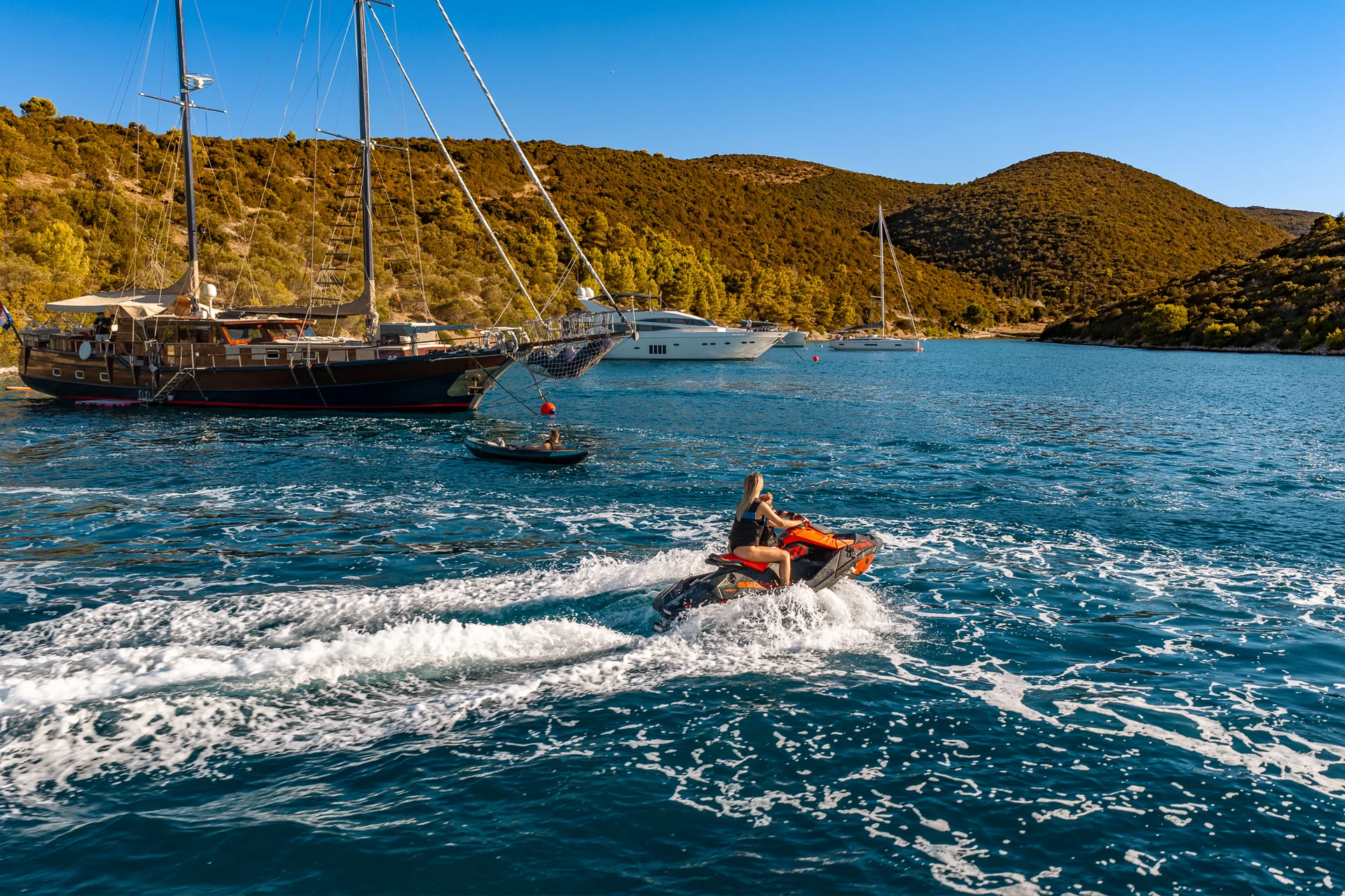 PACHA A person rides a jet ski across a blue bay near several sailboats and yachts anchored off a hilly, wooded coastline.