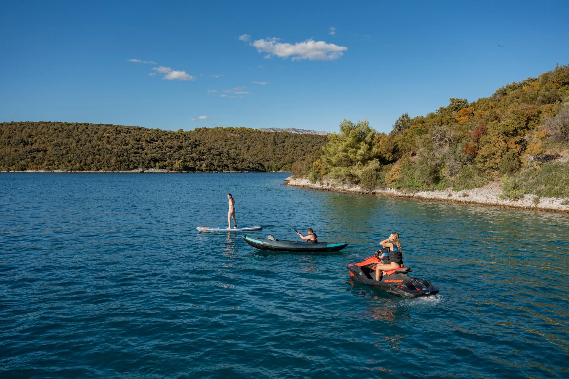 PACHA A person on a stand-up paddleboard, another in an inflatable kayak, and a woman on a jet ski on a calm blue sea near a tree-covered coastline.