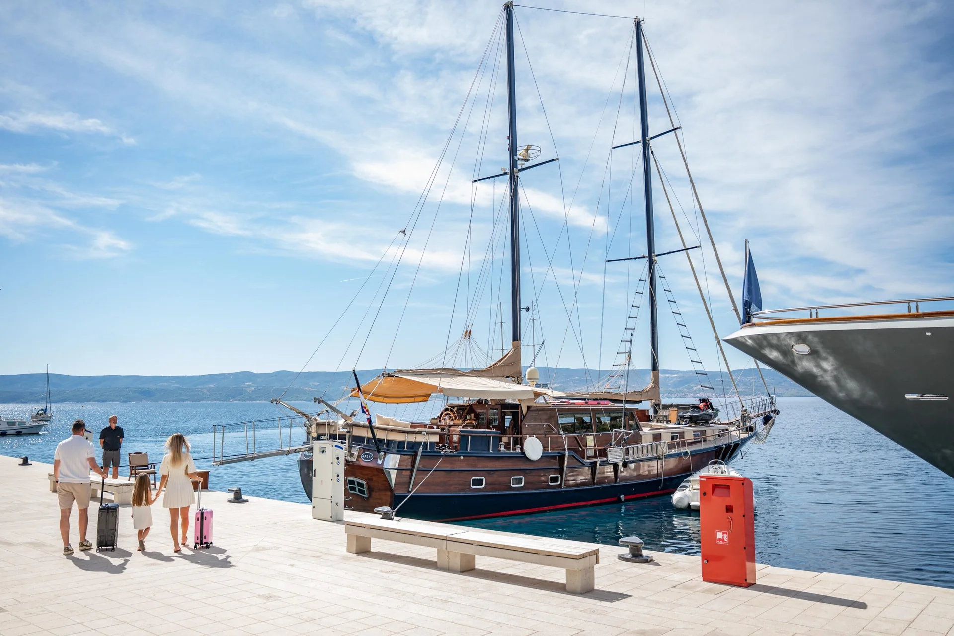 PACHA A family walks along a pier toward a wooden sailboat docked in a harbor.