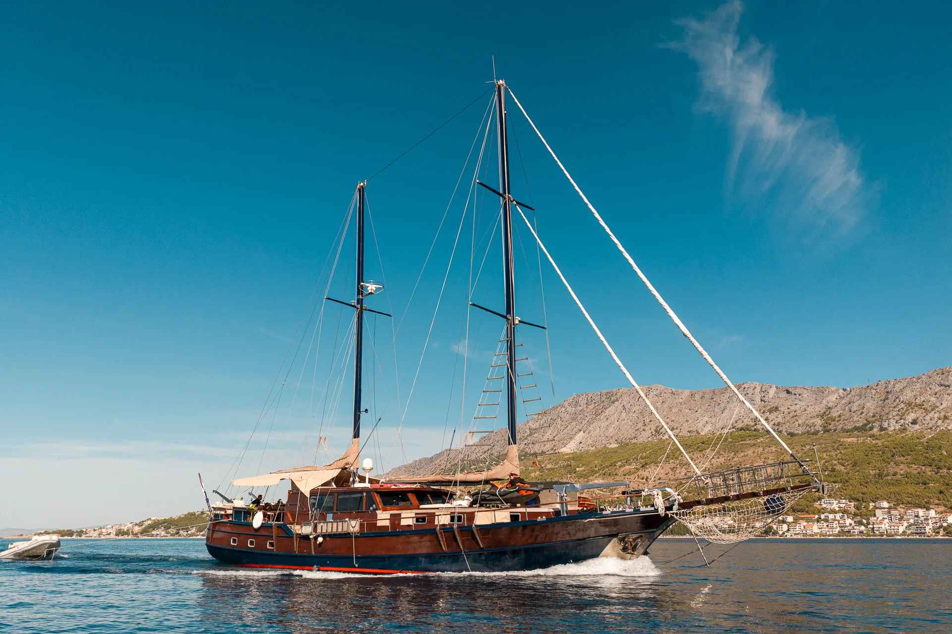 PACHA A wooden sailboat with two masts sailing on the sea with a coastal town and mountains in the background.