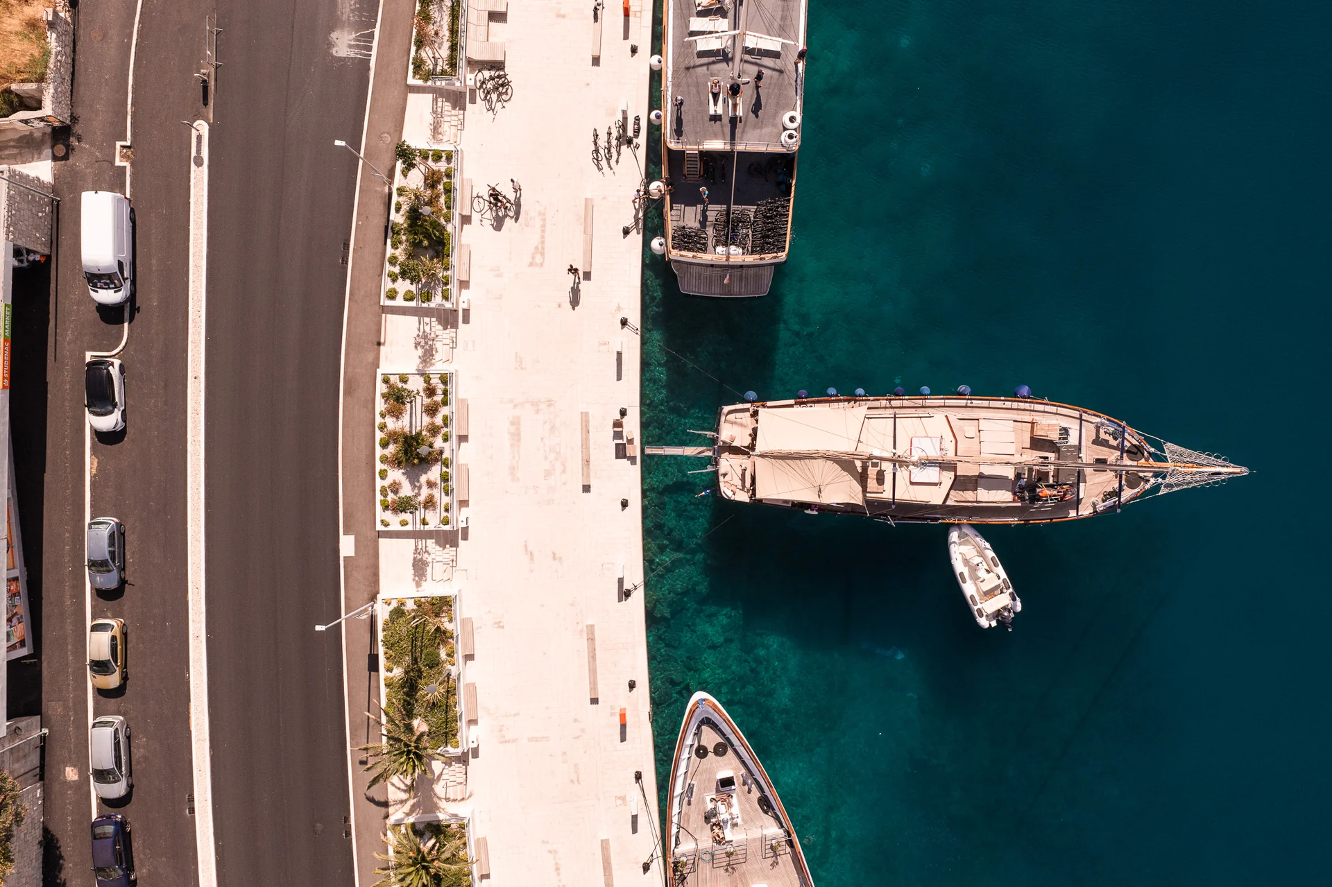PACHA Aerial view of three sailing ships docked along a waterfront promenade next to a road with parked cars.