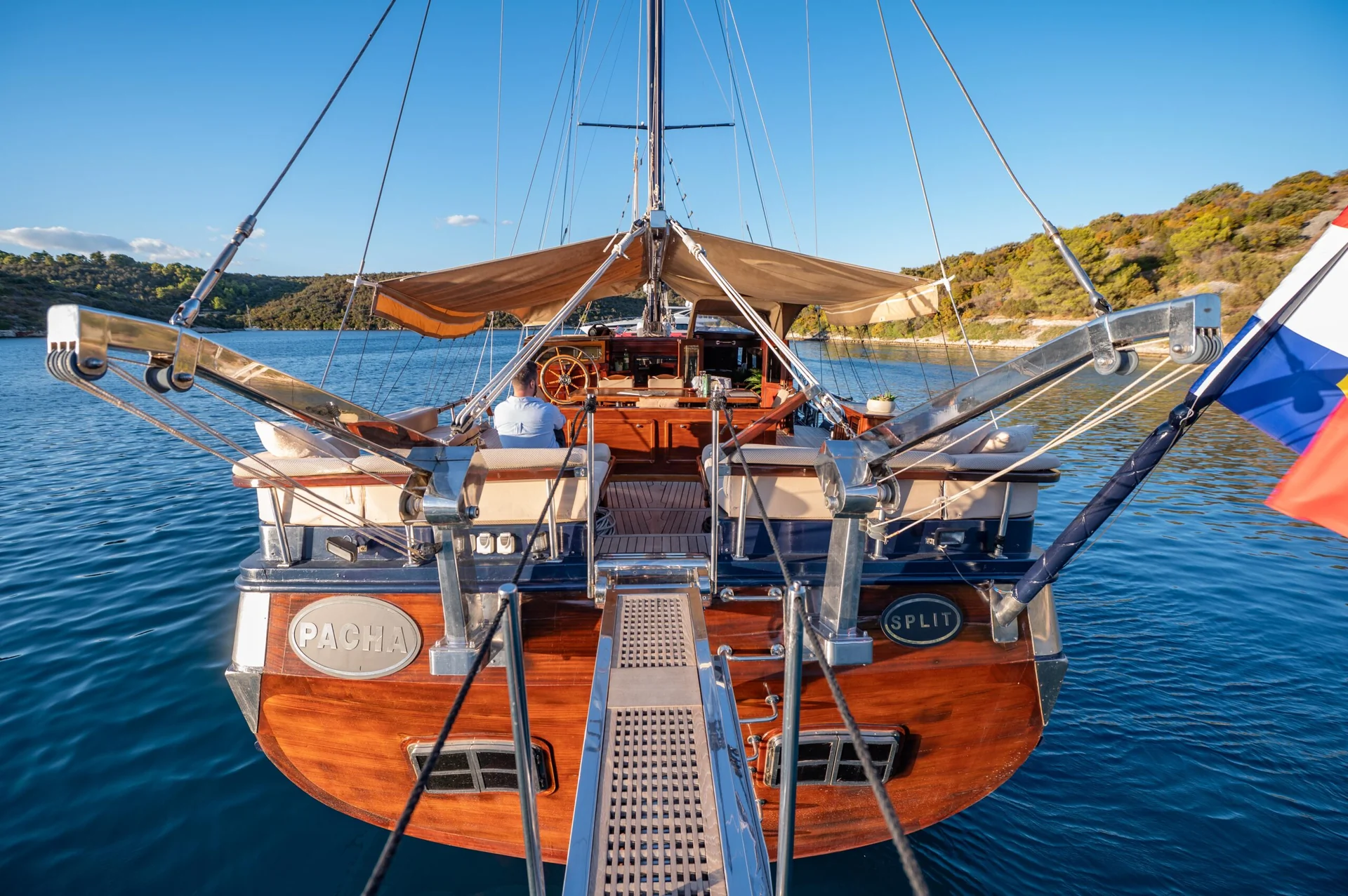 PACHA View from the stern of a wooden sailboat named Pacha, registered in Split, with a person sitting at the helm steering through calm waters.
