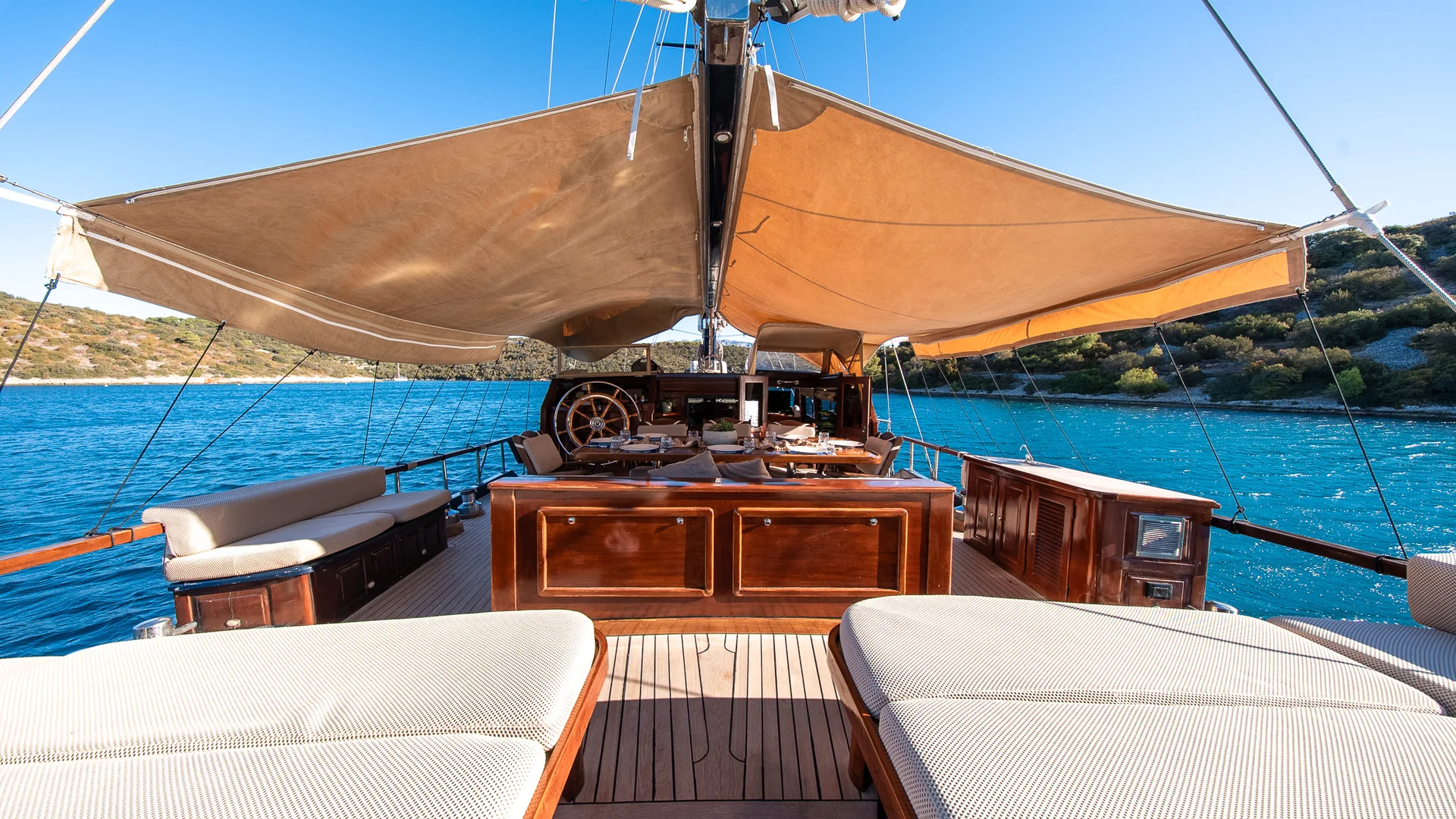 PACHA The deck of a wooden yacht with beige sun awnings, cushioned seating, and an outdoor dining area overlooking the blue sea.