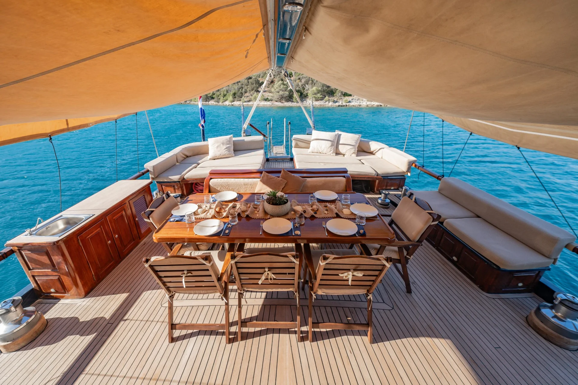 PACHA An outdoor dining table set for six on the teak deck of a yacht, shaded by a tan canopy and overlooking the blue sea.