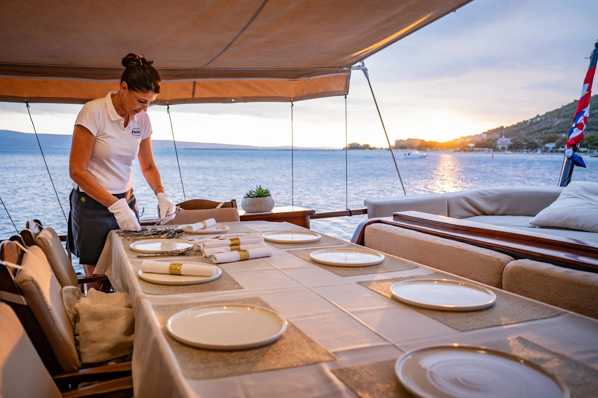 PACHA A crew member in uniform sets a long dining table on the deck of a yacht overlooking the sea at sunset.