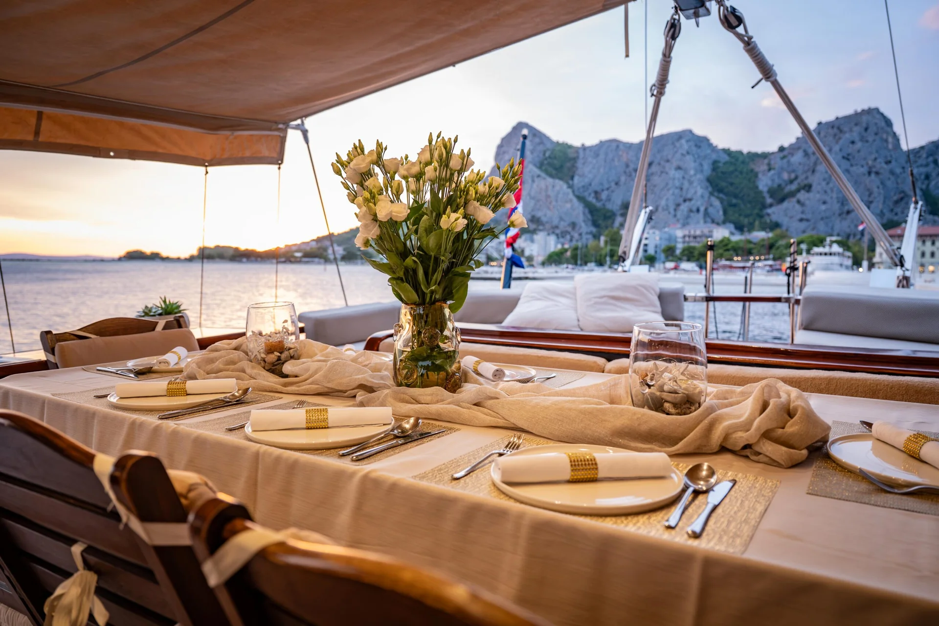 PACHA A dining table set for a meal on the deck of a yacht, featuring a centerpiece of white flowers, with a view of a coastal mountain landscape at sunset.