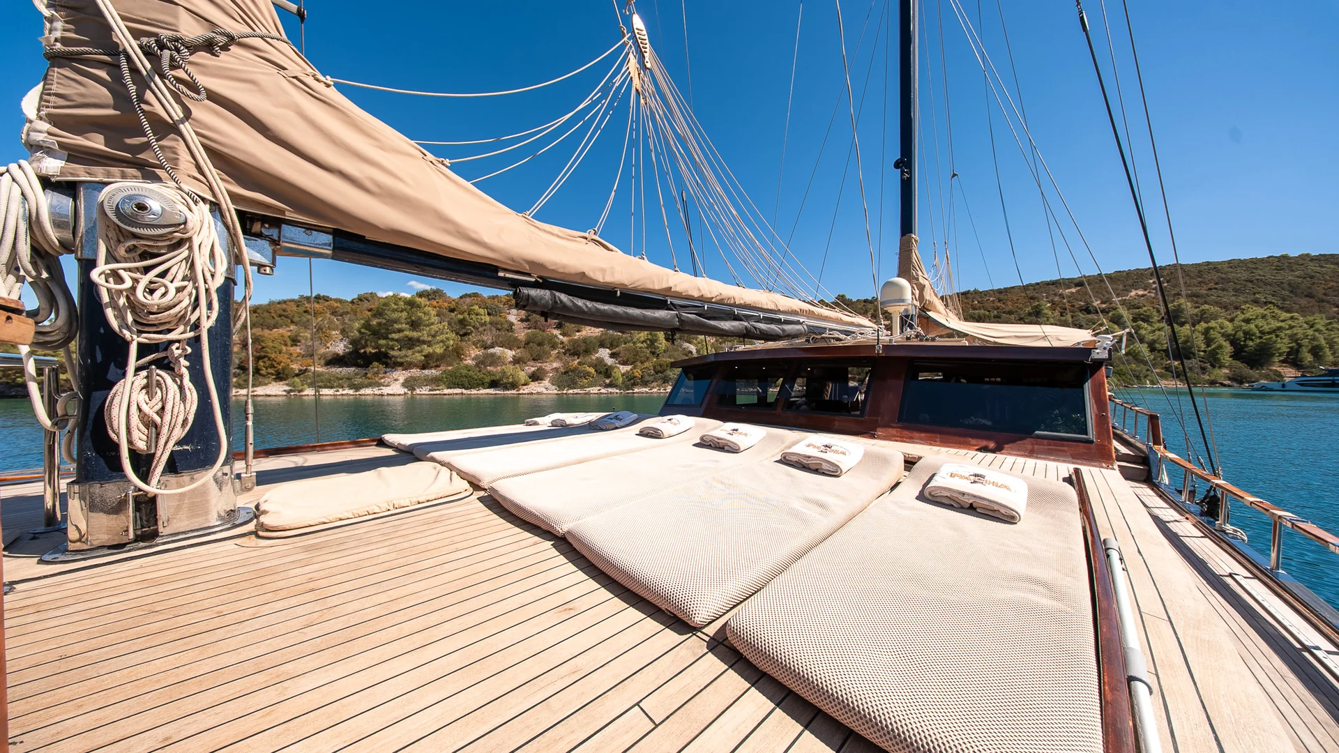 PACHA Wooden deck of a sailboat featuring rows of sun loungers with white towels, looking toward a forested coastline.