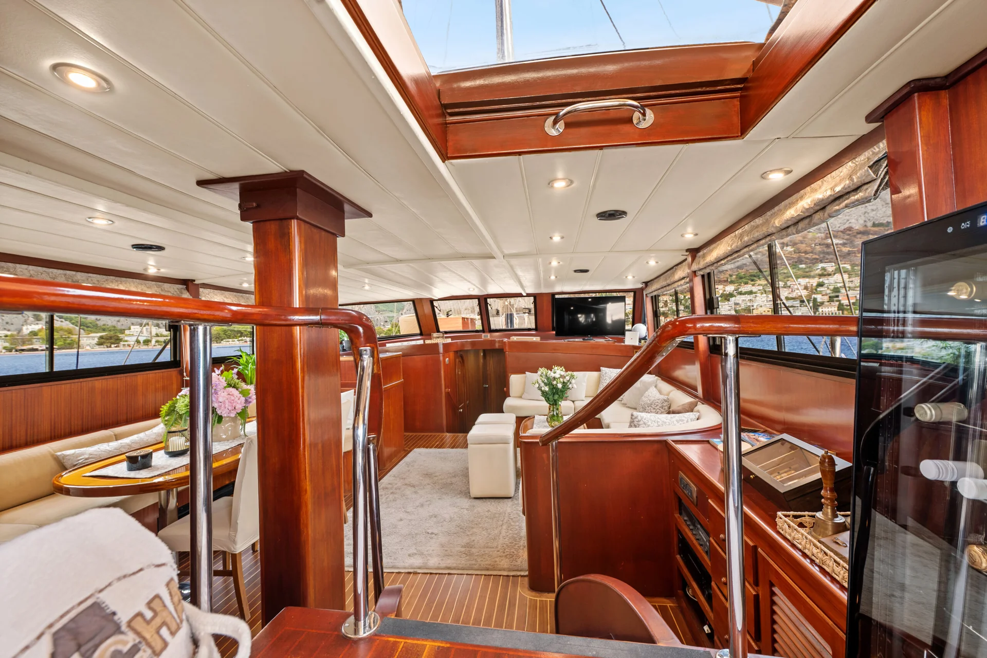PACHA Interior of a wooden yacht cabin featuring a dining area with a circular table and a lounge area with white sofas and a television, viewed from the helm deck.