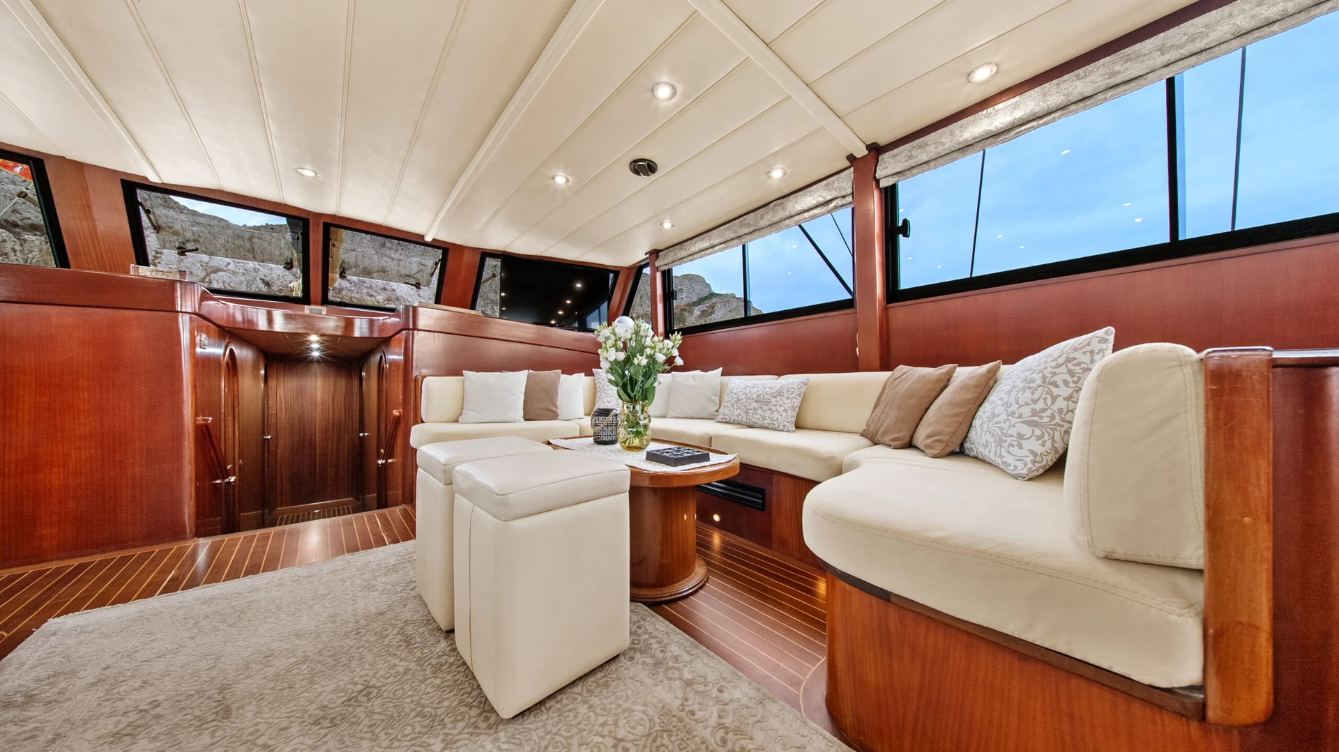 PACHA The interior cabin of a yacht featuring wood-paneled walls, a large cream-colored sectional sofa, two matching ottomans, and a central coffee table with a vase of flowers.