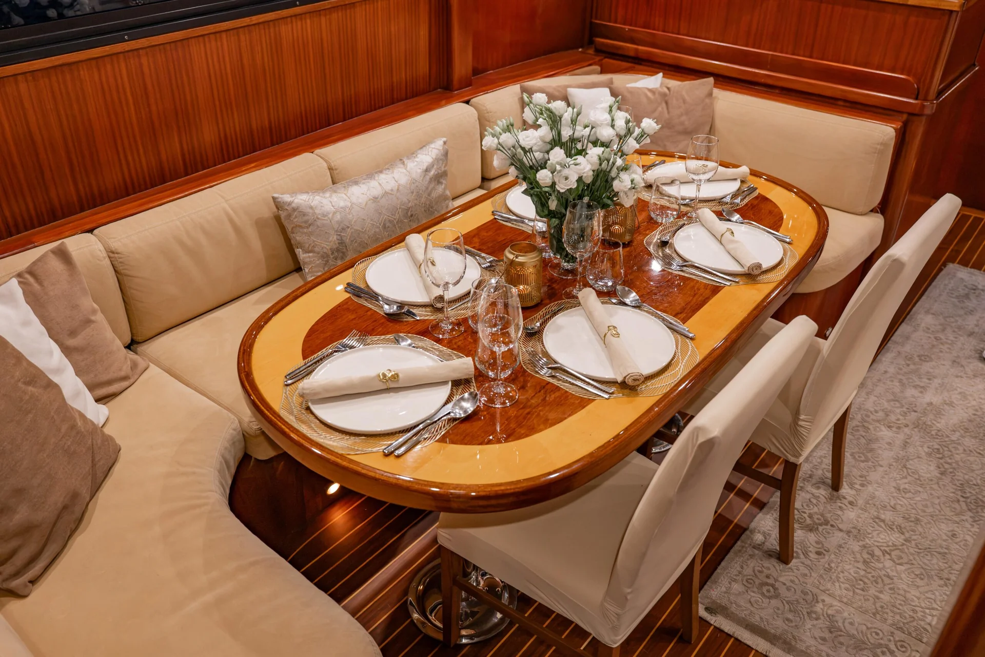 PACHA An oval wooden dining table set for four with white plates, rolled napkins, glassware, and a central bouquet of white flowers, surrounded by cushioned bench seating and chairs.