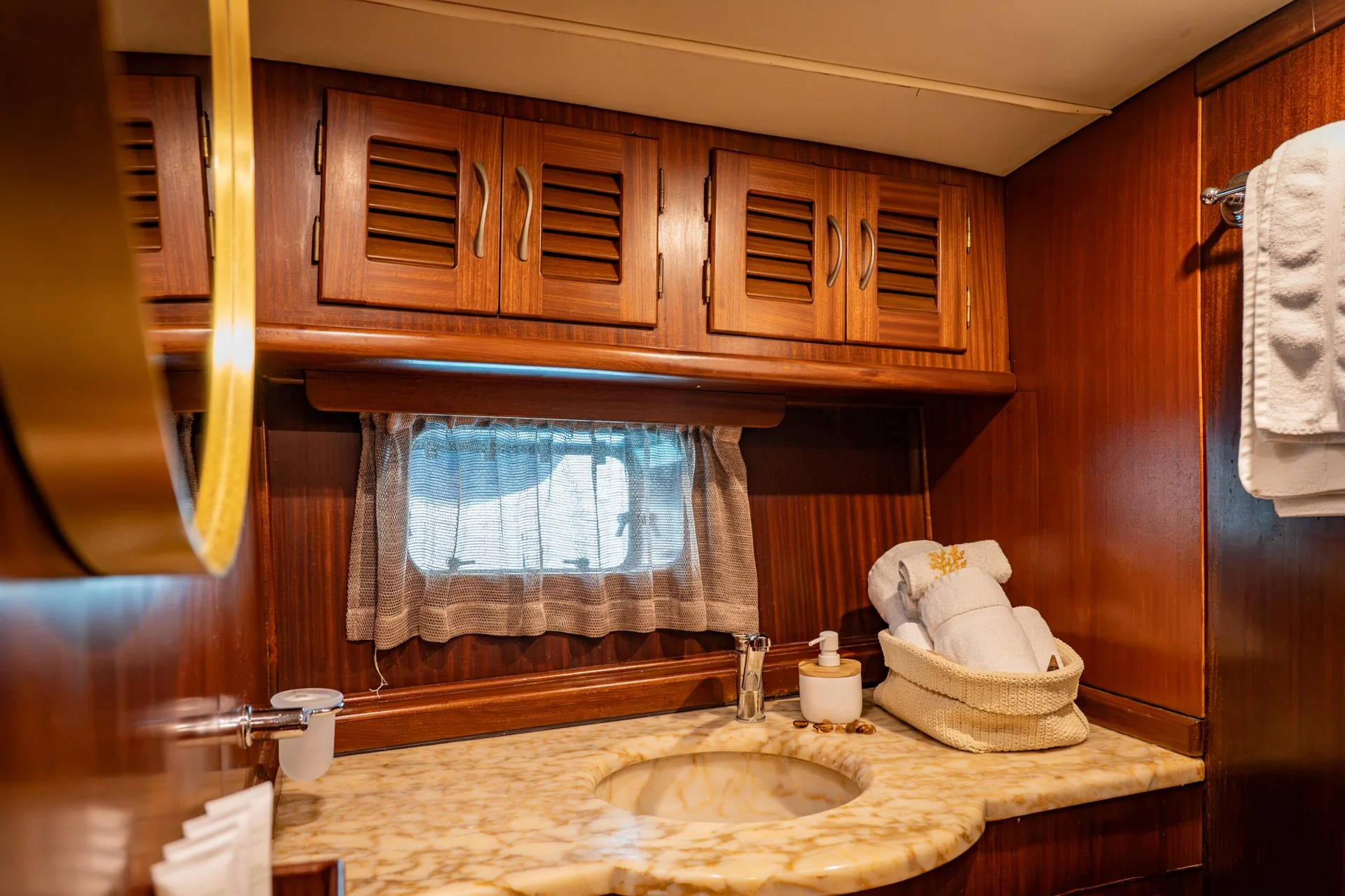 PACHA A boat bathroom with a marble vanity, a white towel basket, wooden louvered cabinets, and a porthole window with a curtain.