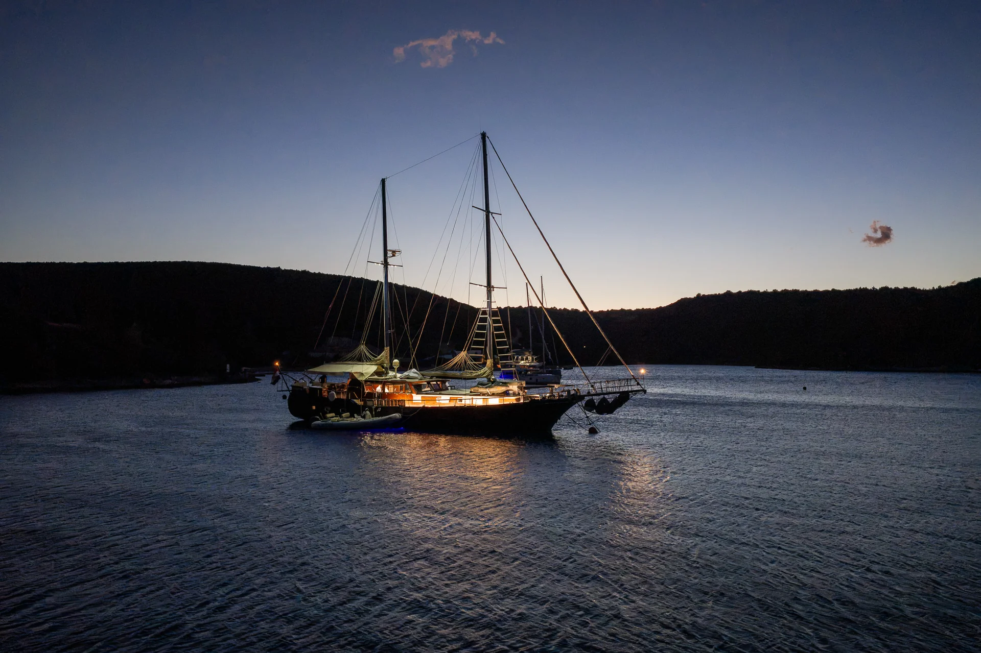 PACHA A sailboat anchored in a calm bay at twilight, with lights illuminated on its deck.