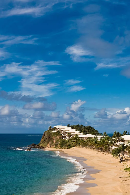 A sandy beach curves along turquoise water, backed by palm trees and modern white buildings atop a small, green cliff under a bright blue sky with scattered clouds.