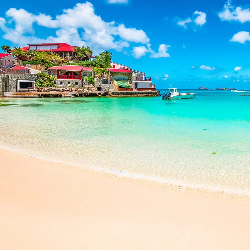 Tropical beach with clear turquoise water, soft sand, and colorful seaside buildings with red roofs under a bright blue sky; a small boat floats near the shore.
