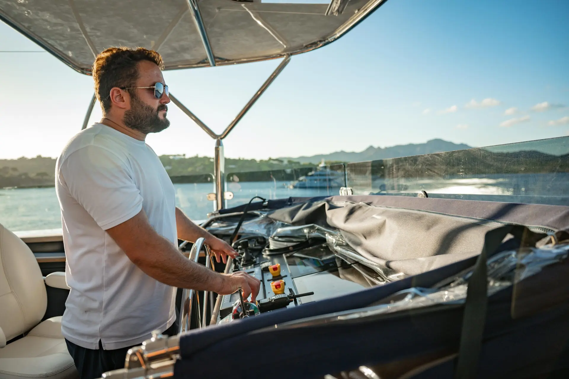 A man wearing sunglasses and a white shirt stands at the helm of a boat, steering on sunny water with distant mountains and another boat visible in the background.