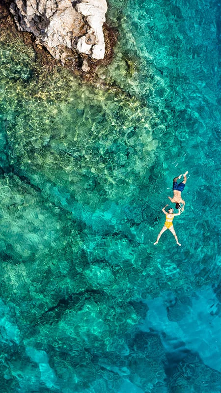 Aerial view of two people swimming in clear turquoise water near rocky coastline, surrounded by vibrant underwater scenery.