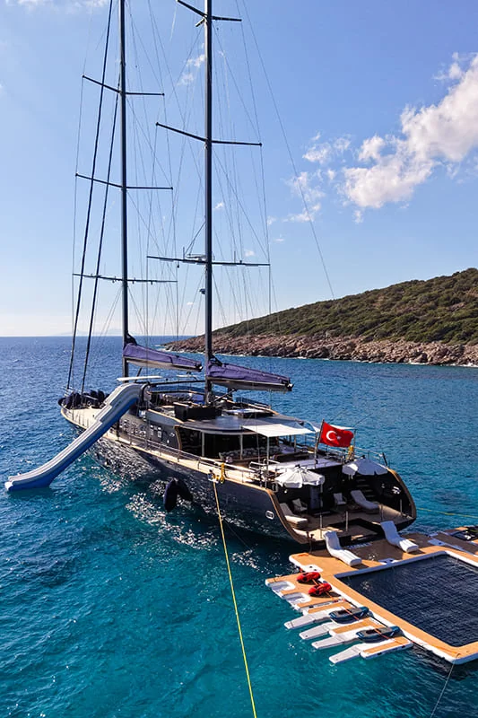 A large yacht with a Turkish flag is anchored in clear blue water near a rocky, green coastline. The yacht has a waterslide and a floating platform with water toys attached to the rear.