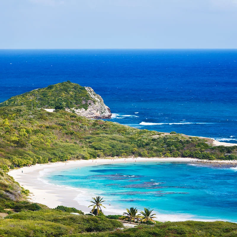 Aerial view of a tropical beach with turquoise waters, white sand, palm trees, and lush greenery, bordered by a rocky hill and deep blue ocean under a partly cloudy sky.
