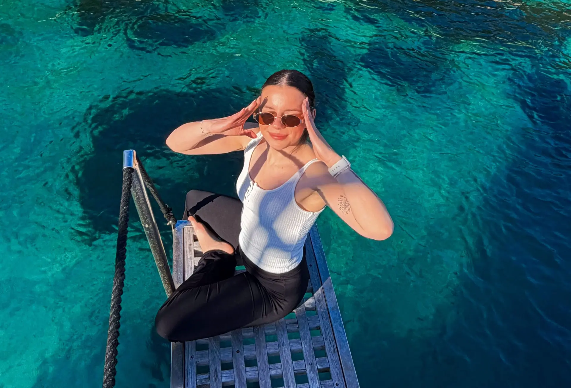 A woman in sunglasses and a white tank top sits on a metal ladder above clear blue water, touching her face with both hands and looking up at the camera, with sunlight casting shadows on her.