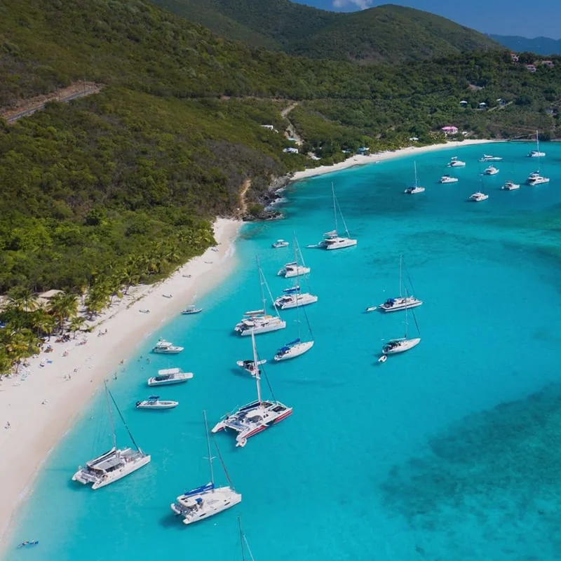 Aerial view of a tropical beach with turquoise water, white sand, several anchored sailboats, and green hills in the background.