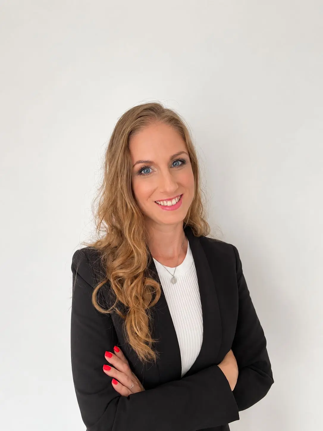 Woman with long wavy blonde hair, wearing a black blazer and white top, smiling with arms crossed in front of a plain white background.