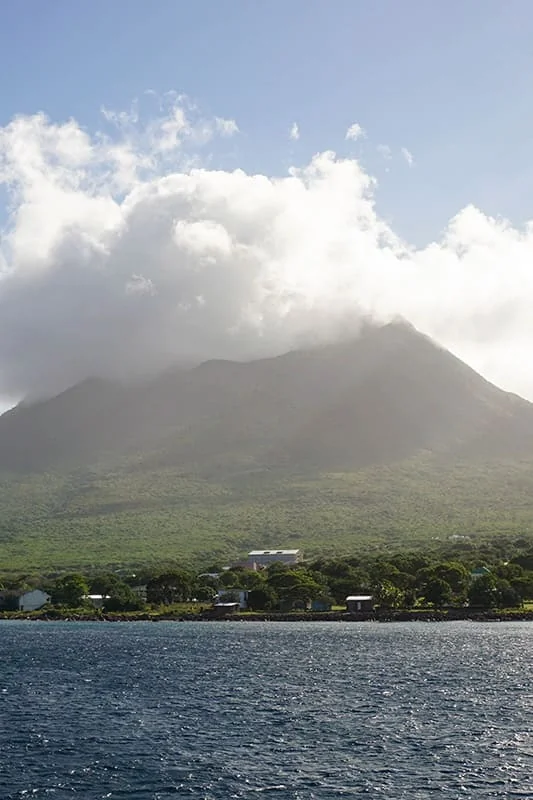 A lush green mountain partly covered by clouds rises behind a small town at the waters edge under a bright blue sky.