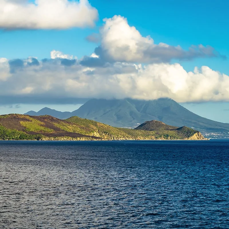 A calm blue sea stretches toward a green, hilly island with a large, cloud-covered mountain in the background under a bright blue sky.