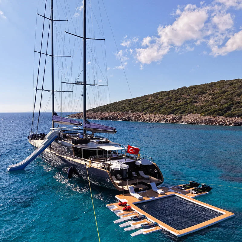 A luxury yacht with a slide and floating deck, anchored in clear blue water near a rocky, green coastline. The yacht displays a Turkish flag.