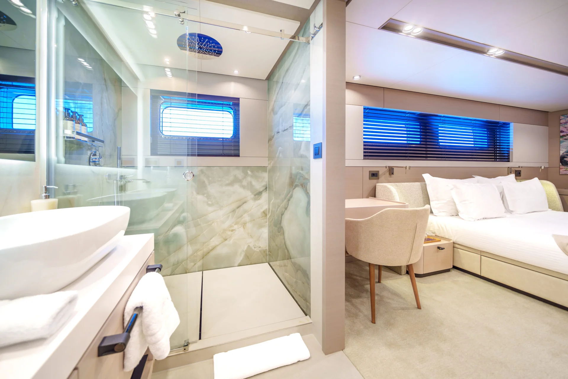 ORIY Modern bathroom with glass shower next to a cozy bedroom featuring a large bed, soft lighting, beige carpet, a desk, and blue-tinted window blinds.
