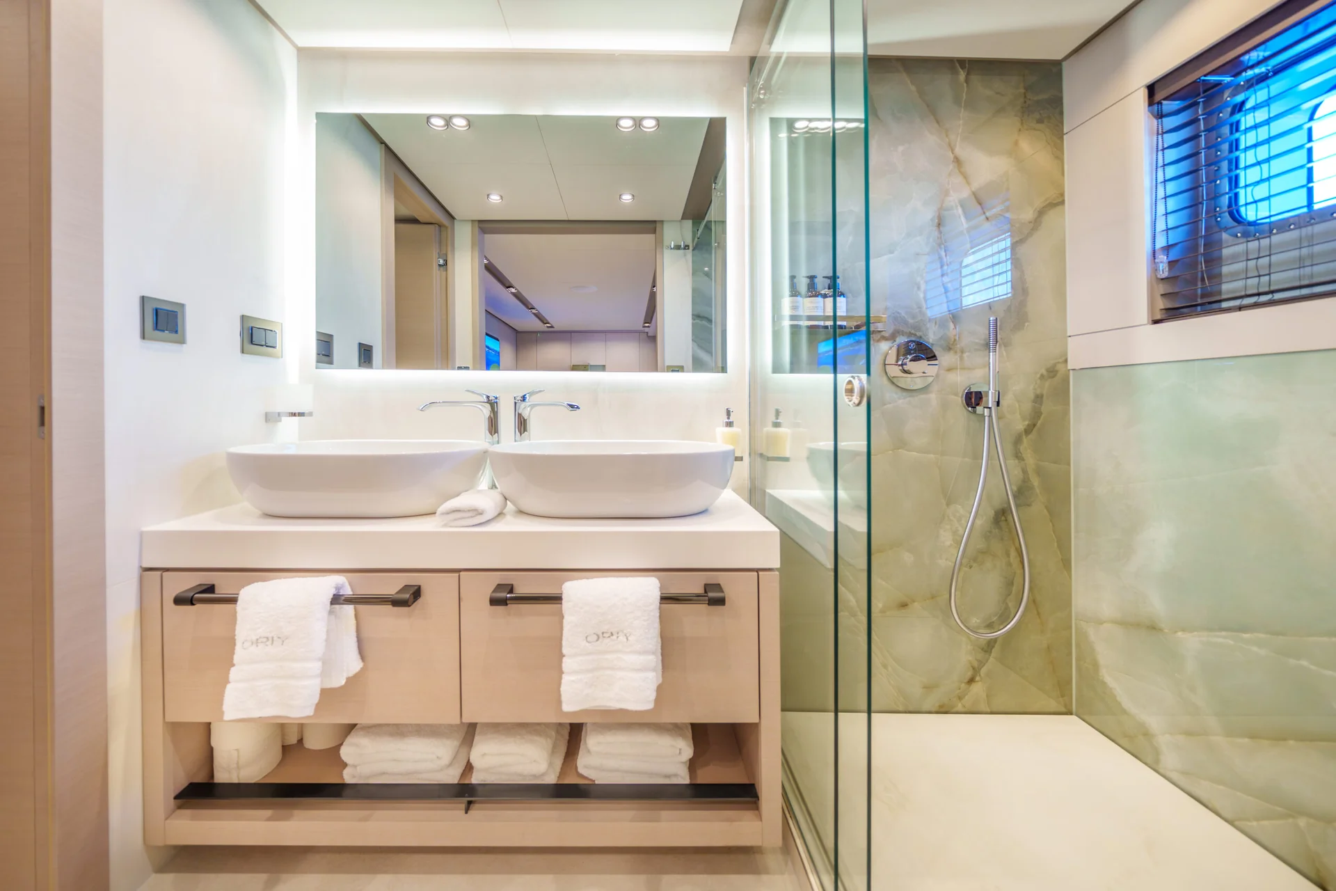 ORIY Modern bathroom with double sinks, towels on shelves, a large mirror, and a glass-enclosed shower. There is a window with blinds above green marble walls next to the shower. Bright, clean, and minimalist design.