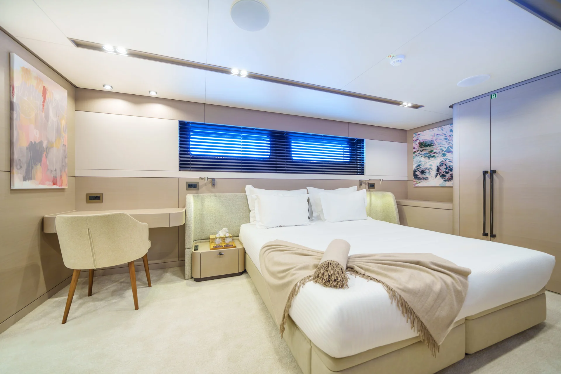 ORIY Modern yacht bedroom with a double bed, beige blanket, and white pillows. Room includes a desk, chair, bedside table, wall art, and window with blinds. The décor is minimalist and bright.
